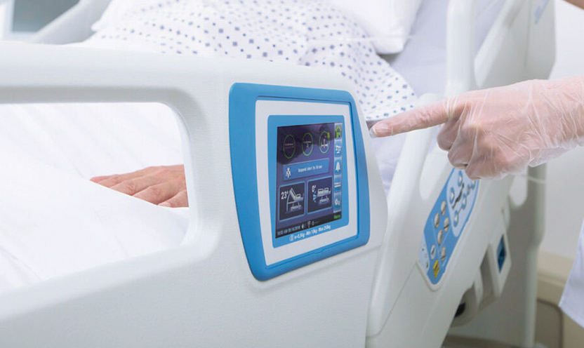 A clinician taps the touchscreen on the Hillrom 900 Accella Smart Bed while a patient rests in bed